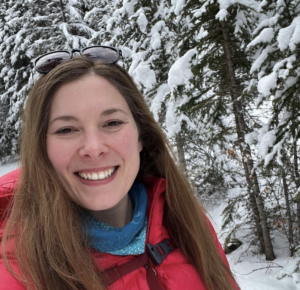 A picture of Laura Kroeker in a red winter jacket smiling in front of a forest covered in snow.